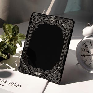 Può includere: Oggetto decorativo rettangolare nero con un bordo argentato ornato di filigrana. L'oggetto ha un centro nero vuoto ed è leggermente angolato. Una sveglia bianca e una piccola pianta sono sullo sfondo. Il testo "FOR TODAY" è visibile su una superficie bianca.