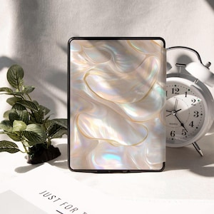 Op de afbeelding: Een zwarte e-reader hoes met een parelmoer, wervelend ontwerp in tinten wit, goud en iriserend. Een witte wekker en een kleine groene plant zijn ook zichtbaar.