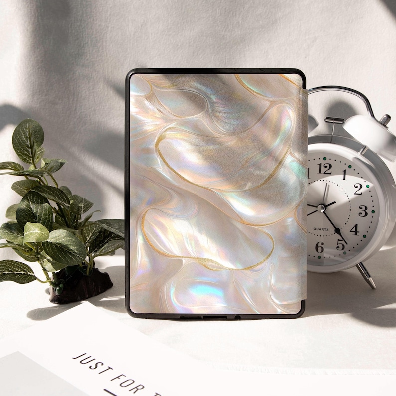 Op de afbeelding: Een zwarte e-reader hoes met een parelmoer, wervelend ontwerp in tinten wit, goud en iriserend. Een witte wekker en een kleine groene plant zijn ook zichtbaar.