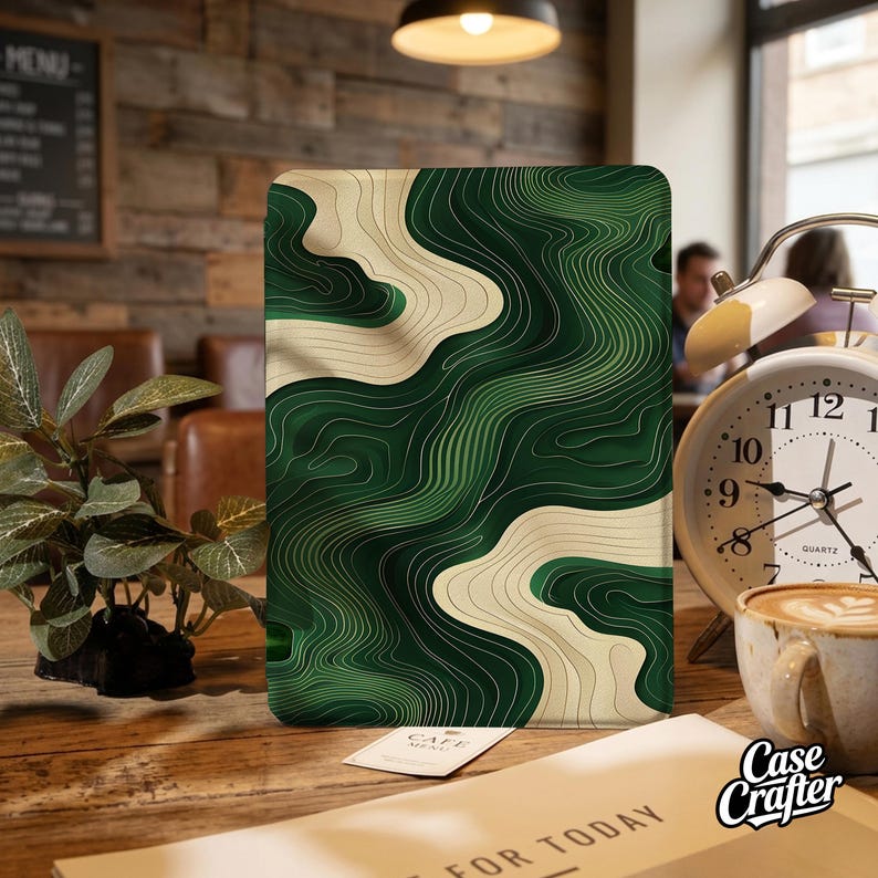 May include: A tablet case featuring a green and cream abstract design. The case has a topographic map-like pattern with curved lines. The Case Crafter logo is visible. The case is on a wooden table with a plant, a clock, and a coffee cup.