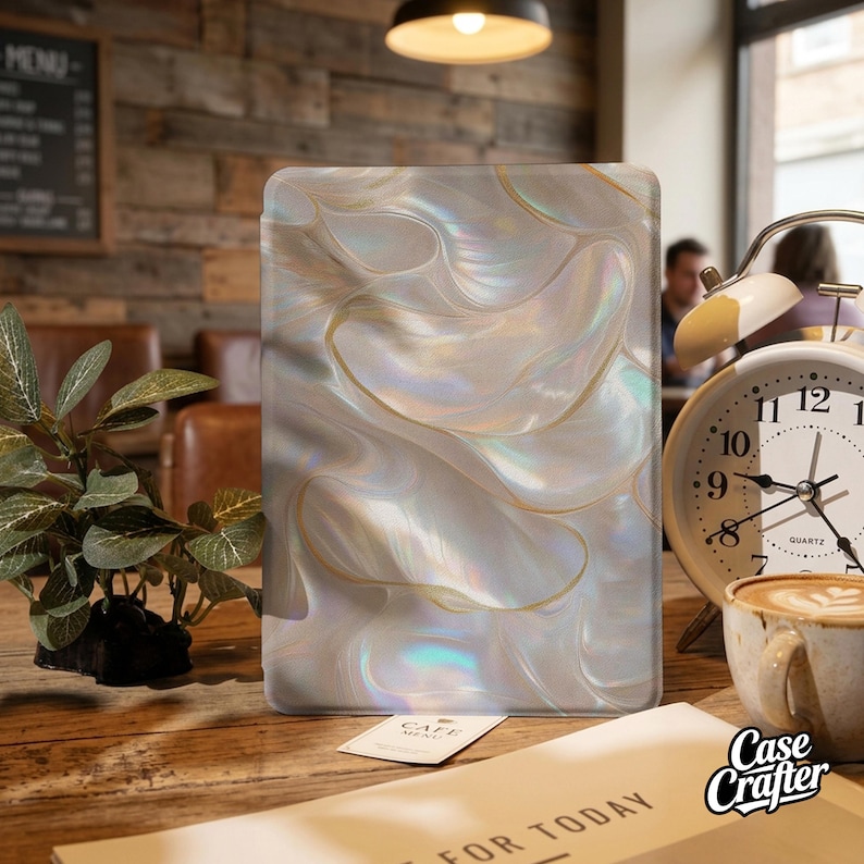 Op de afbeelding: Een rechthoekige tablethoes met een parelmoer, wervelend ontwerp in wit, goud en iriserende tinten. De hoes ligt op een houten tafel met een klok, een kopje koffie en een kleine plant. Het Case Crafter-logo is zichtbaar.