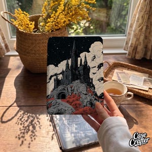May include: A rectangular e-reader cover featuring a detailed black and white illustration of a gothic castle perched on a rocky cliff under a starry night sky, accented with patches of red foliage and white clouds against a dark background.