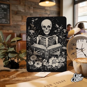 Op de afbeelding: Zwarte tablet hoes met een zwart-witte illustratie van een skelet dat een boek leest, omringd door bloemen. Het Case Crafter logo is zichtbaar. De hoes wordt gepresenteerd op een houten tafel.