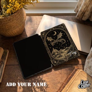 May include: A black e-reader case featuring a gold-coloured illustration of a dragon flying over mountains under a crescent moon and stars. The case has a decorative border and the name Tony written in a script font on the lower right side.