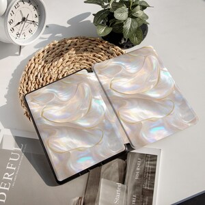 Op de afbeelding: Twee open tabletcases met een parelmoer, wervelend ontwerp in wit, cr&egrave;me en goud. De cases hebben een zwarte rand. Ze liggen op een geweven mat, met een witte klok en een kleine potplant op de achtergrond.