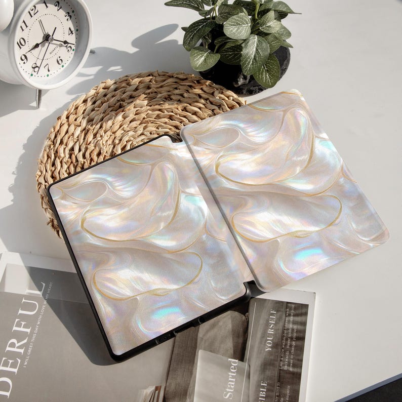 Op de afbeelding: Twee open tabletcases met een parelmoer, wervelend ontwerp in wit, cr&egrave;me en goud. De cases hebben een zwarte rand. Ze liggen op een geweven mat, met een witte klok en een kleine potplant op de achtergrond.