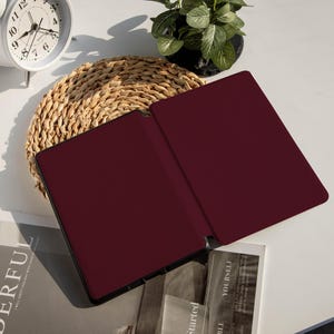 May include: An open, burgundy-colored tablet case with a black border rests on a white surface. The case is next to a woven placemat, a small potted plant, a white analog clock, and a stack of books with the words "YOURSELF" and "Started" visible.