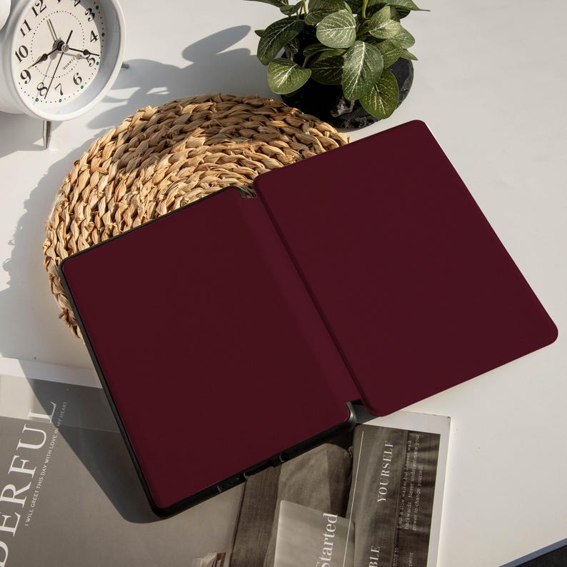 May include: An open, burgundy-colored tablet case with a black border rests on a white surface. The case is next to a woven placemat, a small potted plant, a white analog clock, and a stack of books with the words "YOURSELF" and "Started" visible.