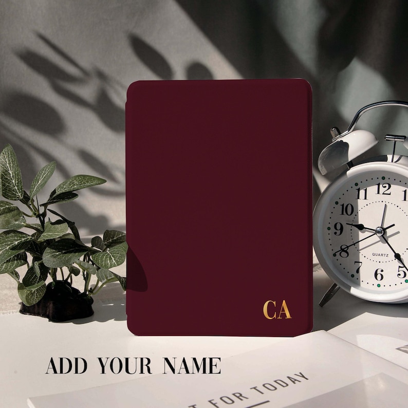 May include: A burgundy tablet case with gold initials "CA" is displayed on a white surface. The text "ADD YOUR NAME" is at the bottom. A white alarm clock and a small plant are also in the image.