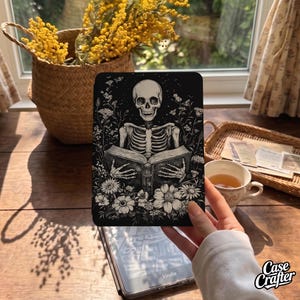 May include: A black e-reader cover featuring a detailed white line art illustration of a human skeleton reading an open book, surrounded by various wildflowers and foliage. The design is monochromatic, showcasing intricate botanical and anatomical details.