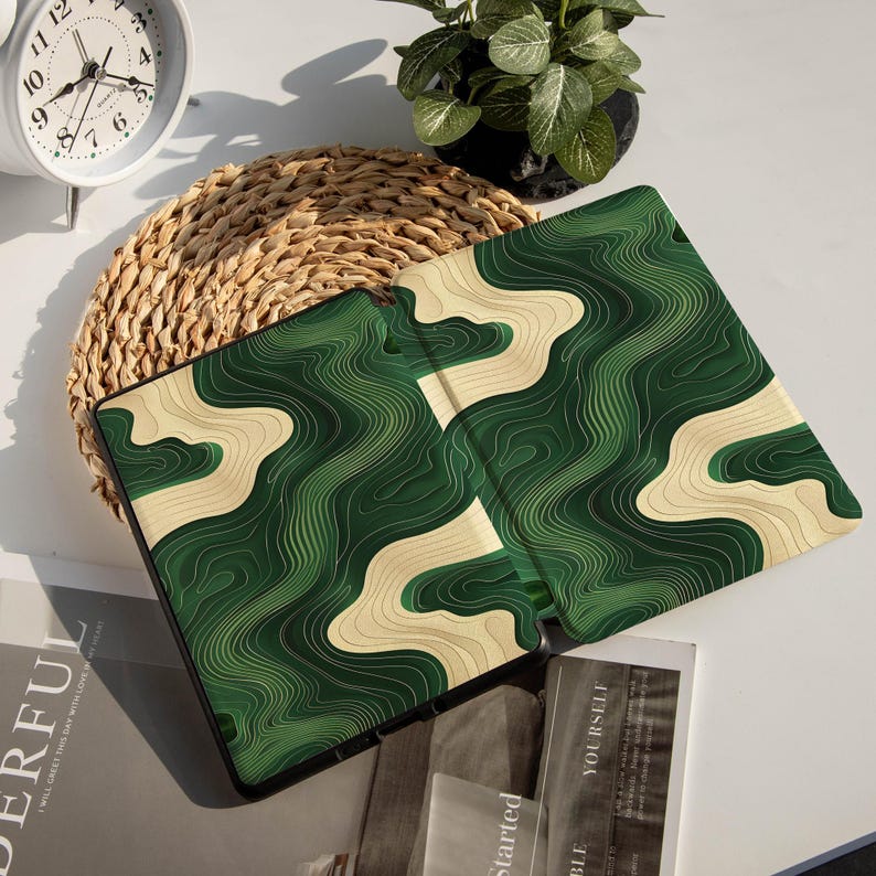 May include: A tablet case featuring a green and beige topographic design. The case is open, displaying the interior pattern. It rests on a white surface, accompanied by a woven mat, a small potted plant, and a clock in the background.