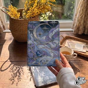 May include: A rectangular e-reader case featuring a stained-glass style design with a crescent moon, stars, and blue roses in shades of purple, blue, and gold. The artwork displays swirling cloud patterns and intricate gold lines throughout the surface.