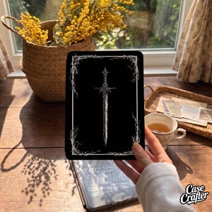 May include: A black e-reader case featuring a white line art illustration of a sword centered within a rectangular frame decorated with thorny vines. The design is printed on a smooth, flat surface, providing a stark contrast between the white graphic and the solid black background of the protective cover.