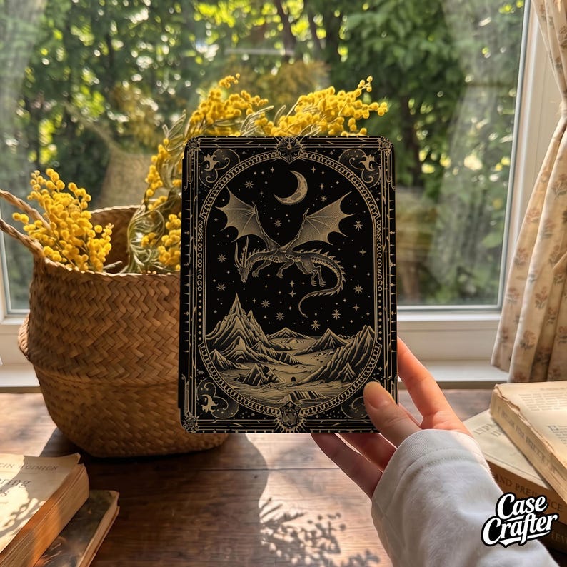 May include: A rectangular black phone case featuring a detailed gold-toned illustration of a dragon flying over a mountain landscape under a crescent moon and stars, all enclosed within an ornate decorative border with intricate filigree patterns.