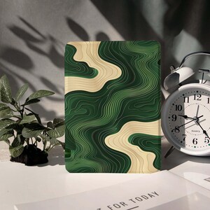 May include: A rectangular case featuring a green and beige topographic design. The case has a layered, wavy pattern in shades of green and beige. The case is on a white surface with a small plant and a white alarm clock in the background.