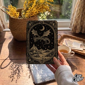May include: A black e-reader cover featuring a gold-toned illustration of a dragon flying over a mountain landscape under a crescent moon and stars, all enclosed within an ornate, decorative border frame with intricate line work and patterns.