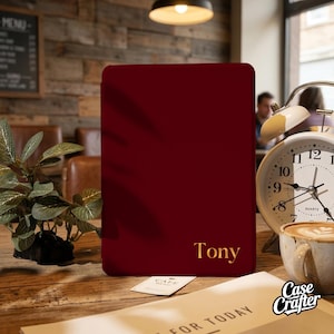 May include: A burgundy phone case with the name "Tony" in gold lettering. The case is rectangular and sits on a wooden table next to a clock and a cup of coffee. A small plant and a menu are also visible.
