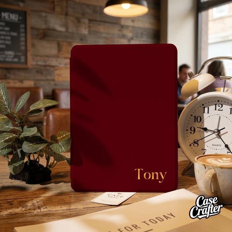 May include: A burgundy phone case with the name "Tony" in gold lettering. The case is rectangular and sits on a wooden table next to a clock and a cup of coffee. A small plant and a menu are also visible.