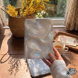 May include: A rectangular e-reader case featuring a pearlescent, iridescent finish with a swirling, liquid-like pattern in shades of white, cream, and soft gold. The surface has a glossy, reflective quality that catches light, creating depth.