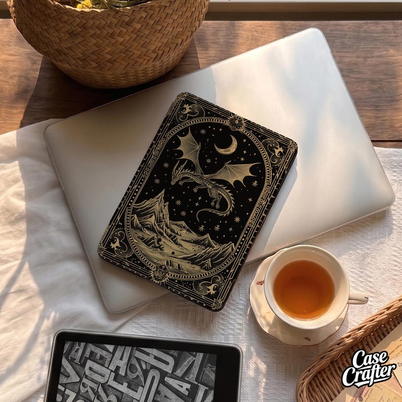 May include: A black e-reader case featuring a gold-coloured illustration of a dragon flying over mountains beneath a crescent moon and stars, enclosed in an ornate decorative border. The case is positioned on a silver laptop next to a white teacup.