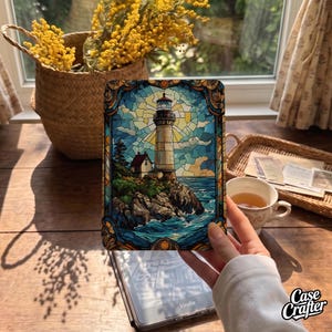 May include: A rectangular e-reader cover featuring a stained glass style illustration of a lighthouse on a rocky cliffside by the sea. The artwork uses shades of blue, yellow, and brown, with a decorative border framing the coastal scene.