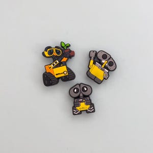 May include: Three colorful robot-themed charms. One is yellow with a plant, another is yellow and gray, and the third is gray with the word "WALL" on it. These charms are likely for shoes or accessories.