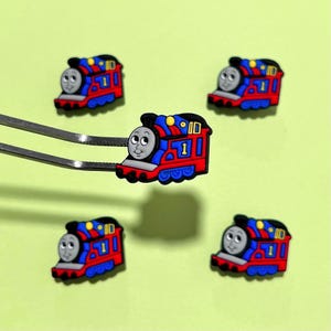 May include: Five colorful train charms with a smiling face, red body, blue accents, and the number 1. One charm is held by silver tweezers against a light green background. The train charms are likely for shoes or accessories.
