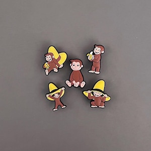 May include: Five brown monkey-shaped charms with black outlines. Two monkeys wear yellow hats, two hold bananas, and one sits. The charms are on a gray background.