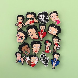 May include: A collection of colorful Betty Boop character charms in various poses. The charms feature the iconic cartoon character in different outfits and expressions, set against a light green background.