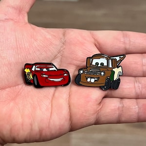 May include: Two enamel pins featuring characters from the animated movie Cars. One pin is a red race car with the number 95 and a flame design. The other is a brown tow truck with a friendly face.