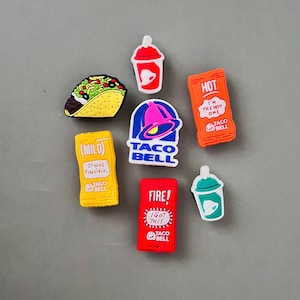May include: A collection of colorful Taco Bell-themed charms. The charms include a taco, drink cups, sauce packets with "HOT", "MILD", and "FIRE!" text, and the Taco Bell logo. These charms are likely for shoes or accessories.
