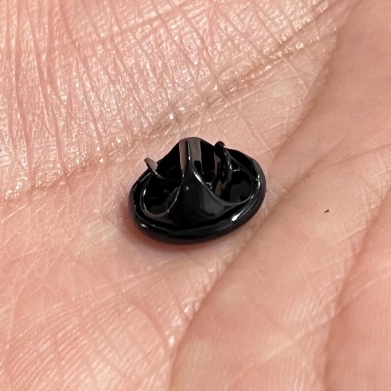 K&ouml;nnte beinhalten: Kleiner, schwarzer Ansteckpin mit rundem Sockel und erhabenem, strukturiertem Design. Der Pin hat eine gl&auml;nzende, reflektierende Oberfl&auml;che und einen Stift zur Befestigung. Der Pin ist wahrscheinlich aus Metall und wird an Kleidung oder Accessoires befestigt.