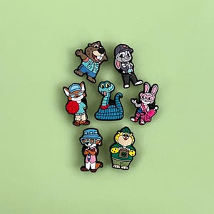 May include: A collection of seven colorful cartoon character charms. The charms feature various animals, including a beaver, rabbit, fox, snake, and bear, each wearing clothing and accessories. The charms are set against a solid green background.