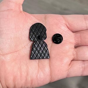 K&ouml;nnte beinhalten: Ein schwarzer Emaille-Pin in Form einer Person mit Bob-Frisur, die ein Kleid mit einem wei&szlig;-schwarzen Karomuster tr&auml;gt. Ein separater schwarzer Pin-R&uuml;cken befindet sich neben dem Pin.