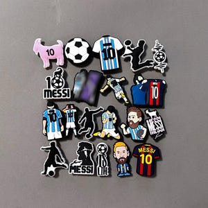 May include: A collection of colorful shoe charms featuring soccer-themed designs. The charms include soccer balls, jerseys with the number 10, and images of a soccer player. Some charms have the word "MESSI" on them.
