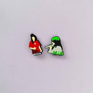 May include: Two shoe charms featuring a singer. One charm shows a figure in a red shirt with the name "Billie" on the bottom. The other charm depicts a figure with green hair holding a microphone, with the word "Billie" in green.