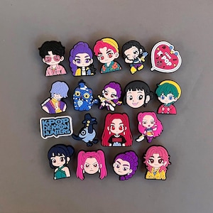 May include: A collection of nineteen colorful rubber shoe charms featuring various chibi-style characters, a blue patterned creature, a black bird, and two heart-shaped icons with the text I LOVE K-POP and K-POP DEMON HUNTERS in bold lettering.