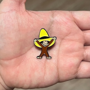 May include: A colorful enamel pin featuring a cartoon monkey wearing a yellow sombrero. The monkey is brown with black outlines, holding the hat with both hands. The pin has a black border.