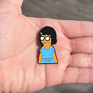 K&ouml;nnte beinhalten: Ein Cartoon-Pin mit einer Figur mit schwarzen Haaren, Brille und einem blauen Oberteil. Das Oberteil tr&auml;gt den Text "EVERYTHING IS FINE". Der Pin ist eine lebendige Mischung aus Farben, darunter Schwarz, Blau, Gelb und Wei&szlig;.