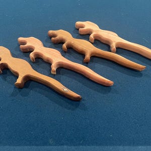 May include: Four wooden dinosaur-shaped objects are arranged diagonally on a blue surface. The dinosaurs are light brown with visible wood grain and have a stylized design. They appear to be decorative or functional items.