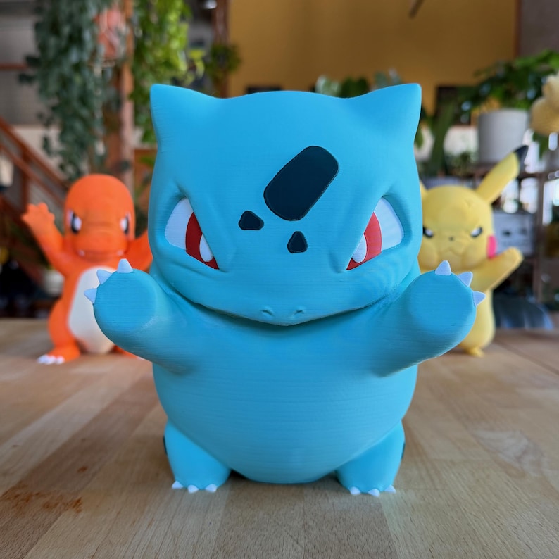 Puede incluir: Figura de Bulbasaur azul claro con ojos rojos y detalles negros. La figura est&aacute; de pie con los brazos extendidos. Otras figuras de Pok&eacute;mon, incluyendo Charmander y Pikachu, son visibles en el fondo.