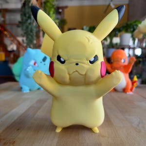 Puede incluir: Una figura amarilla de Pikachu con orejas con puntas negras, marcas rojas en las mejillas y una expresi&oacute;n enfadada. Otras figuras de Pok&eacute;mon, incluyendo Bulbasaur y Charmander, son visibles en el fondo. Las figuras est&aacute;n sobre una superficie de madera.
