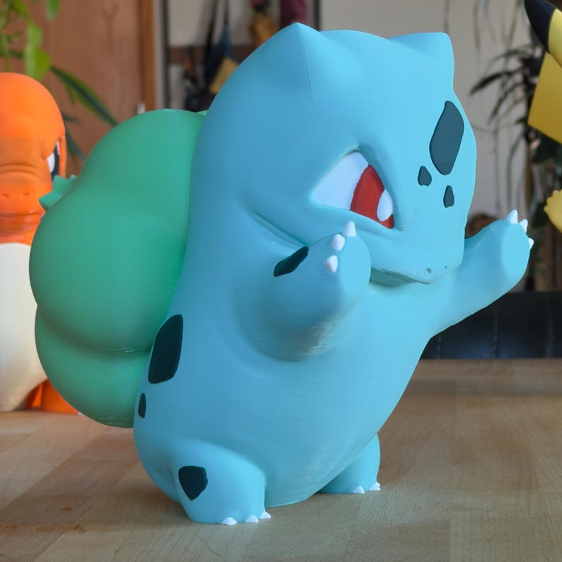 Puede incluir: Figura de Bulbasaur azul claro con un bulbo verde en la espalda. La figura tiene manchas negras, ojos rojos y garras blancas. Bulbasaur est&aacute; posando con los brazos levantados. Otras figuras son visibles en el fondo.