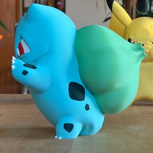 Puede incluir: Figura de Bulbasaur azul claro con manchas negras y un bulbo verde en la espalda. La figura est&aacute; sobre una superficie de madera. Una figura de Pikachu amarilla es parcialmente visible en el fondo.