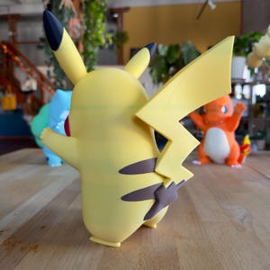 Puede incluir: Una figura amarilla de Pikachu con orejas con puntas negras y una cola en forma de rayo. La figura tiene marcas marrones y una mejilla roja. Otras figuras de Pok&eacute;mon son visibles en el fondo, incluyendo un Bulbasaur azul y un Charmander naranja.