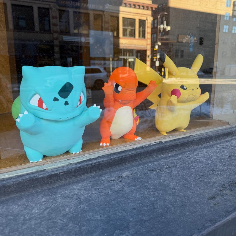 Puede incluir: Tres coloridas figuras de Pok&eacute;mon se exhiben en un escaparate. Un Bulbasaur azul claro, un Charmander naranja y un Pikachu amarillo posan con los brazos levantados. Las figuras est&aacute;n frente a una ventana con una calle de la ciudad visible al fondo.