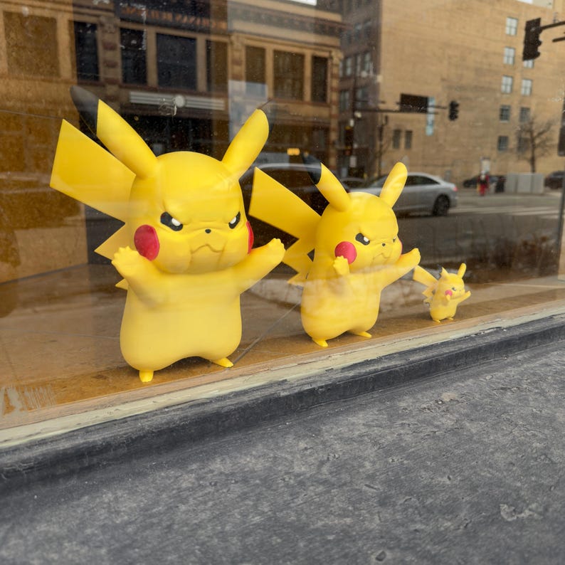 Puede incluir: Tres figuras amarillas de Pikachu con marcas rojas en las mejillas y orejas con puntas negras se exhiben en un escaparate. Las figuras son de diferentes tama&ntilde;os, la m&aacute;s grande en la parte delantera y la m&aacute;s peque&ntilde;a en la parte trasera. El fondo muestra una calle de la ciudad.