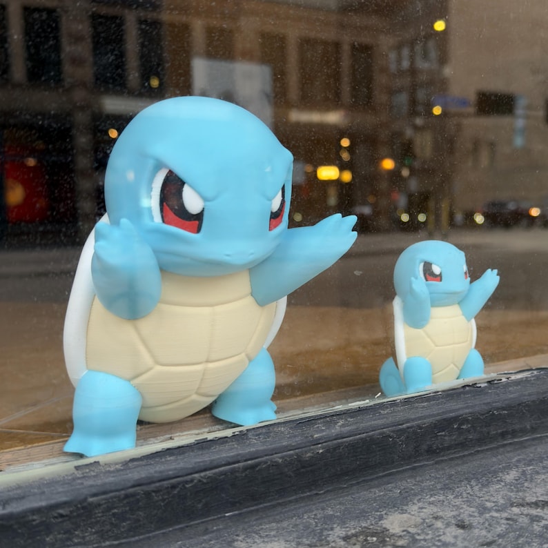Puede incluir: Se muestran dos figuras azules de Squirtle, una grande y otra peque&ntilde;a. La figura m&aacute;s grande tiene los brazos levantados, mientras que la m&aacute;s peque&ntilde;a tiene los brazos cerca de la cabeza. Ambas tienen caparazones beige y ojos rojos.