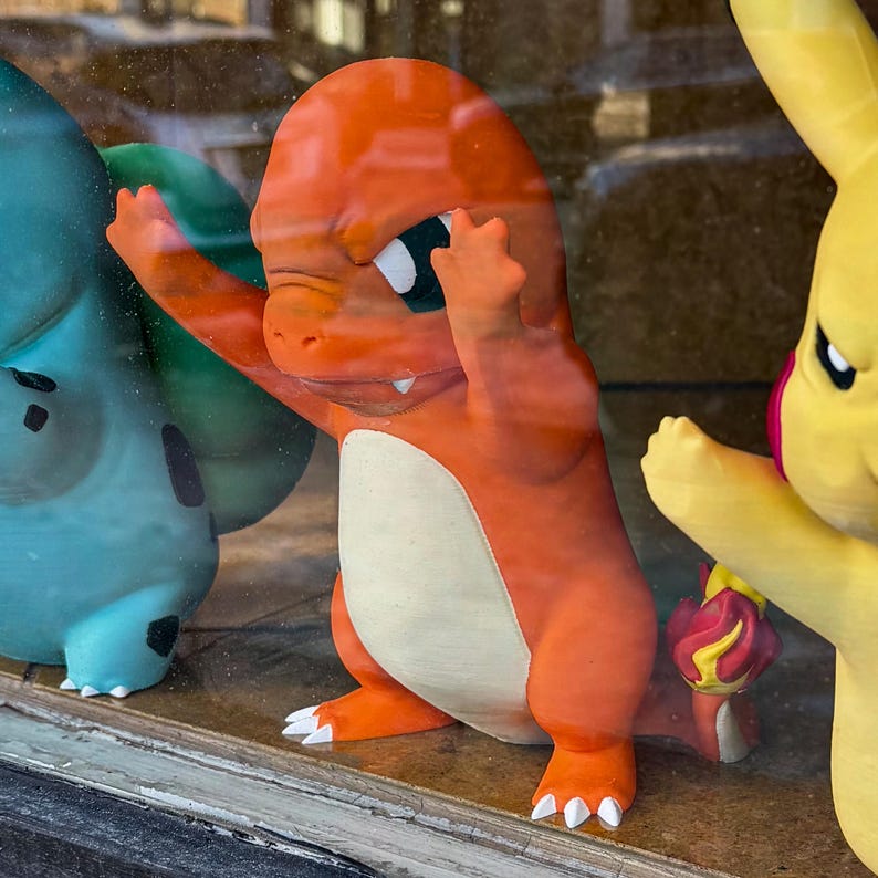 Puede incluir: Una figura de Charmander naranja con los brazos levantados, junto a un Bulbasaur verde y una figura de Pikachu amarilla. El Charmander tiene la barriga blanca y ojos negros. Las figuras se exhiben en un escaparate.