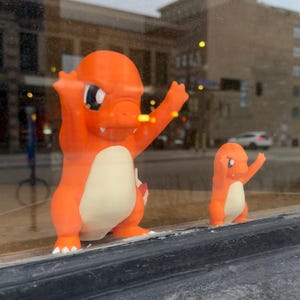 Puede incluir: Dos figuras de Charmander naranjas, una grande y otra peque&ntilde;a, est&aacute;n una al lado de la otra. La figura m&aacute;s grande tiene los brazos levantados, mientras que la m&aacute;s peque&ntilde;a imita la pose. Ambas tienen vientres color crema y se exhiben en una ventana.